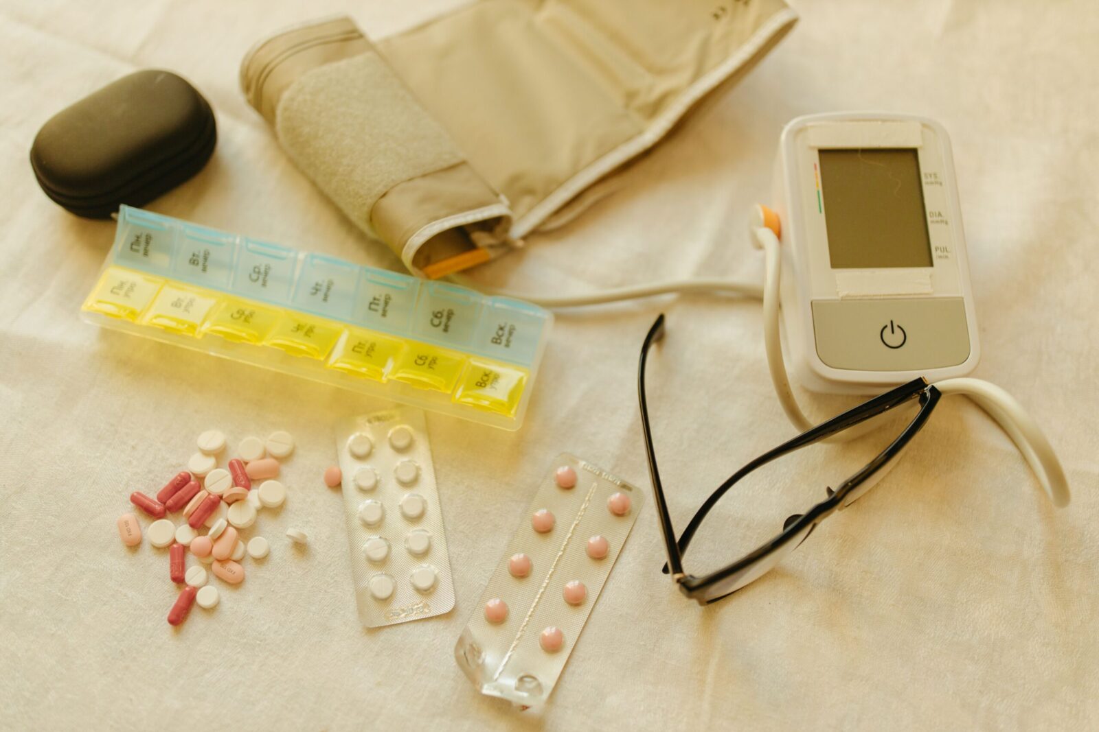 Flat lay image of healthcare essentials including a blood pressure monitor, pills, and eyeglasses on a white surface.